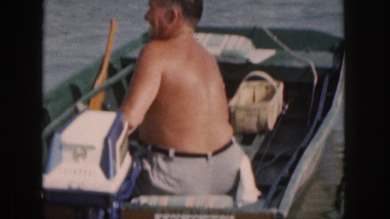 In 1961 Michigan home-movie footage, a shirtless man sits in a small motorboat near a blue-and-white outboard motor and wooden oar.