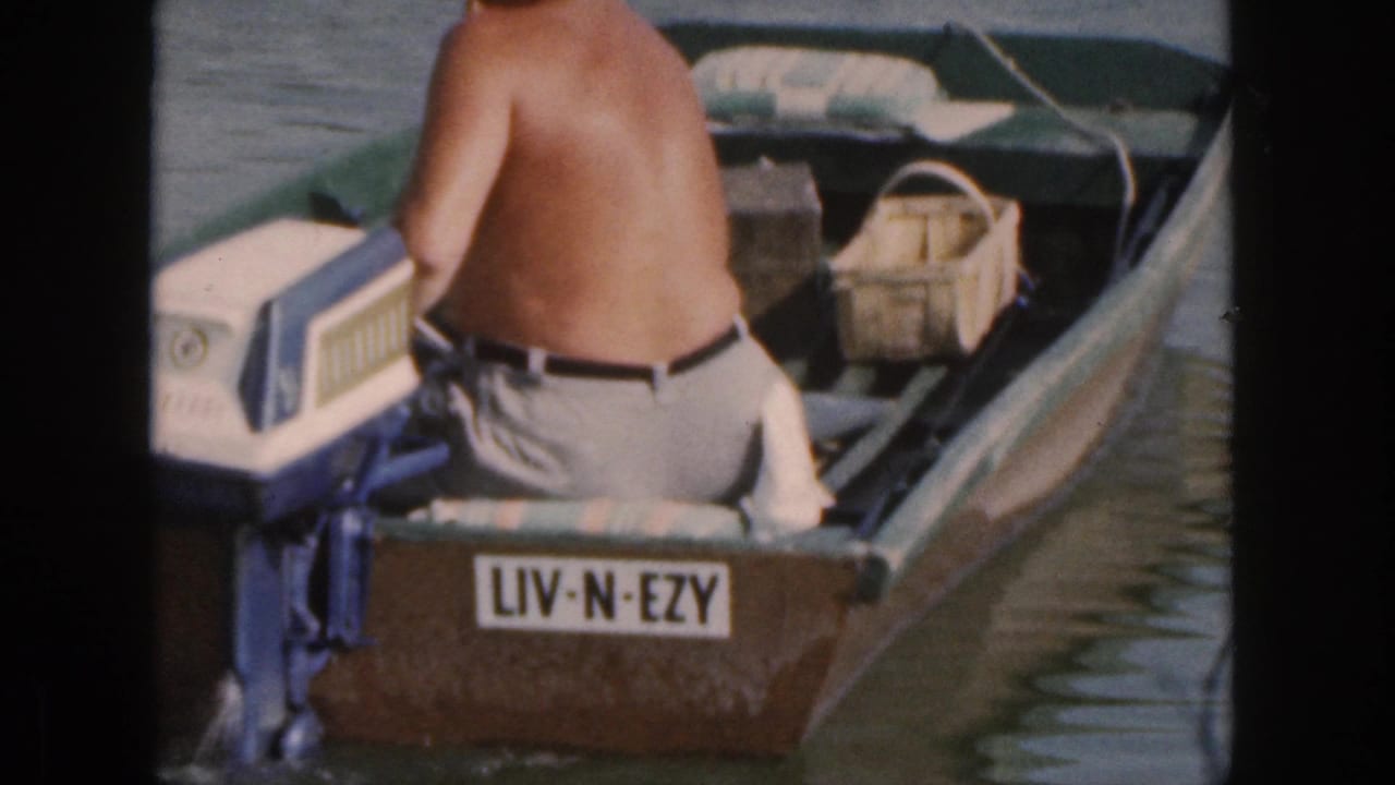 A shirtless man faces away in a small Michigan motorboat, with the stern plate reading LIV-N-EZY and a basket visible inside.