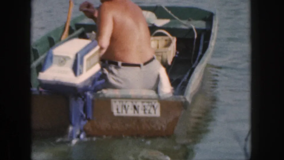 The man leans forward in the small 1961 Michigan motorboat, with the blue-and-white motor, stern plate, and water around the boat visible.