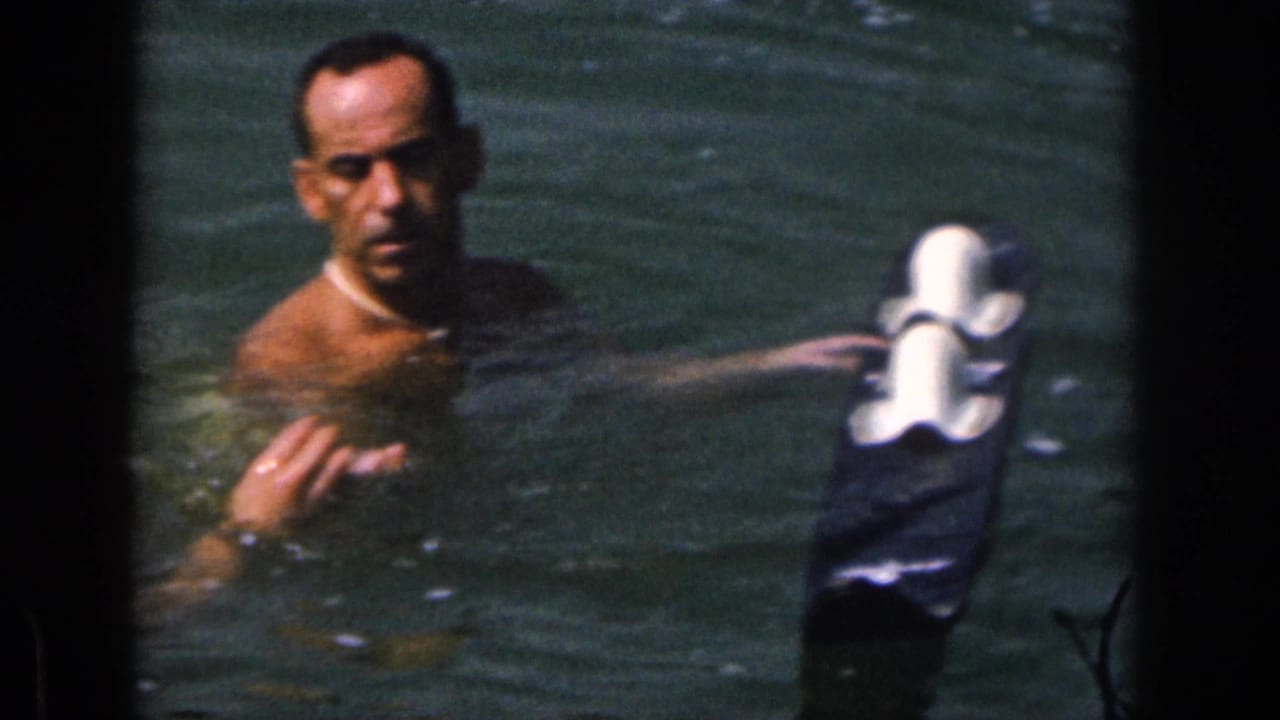 Adult man in Michigan lake water in 1961, holding near a pair of white-topped water skis in soft 8mm footage.
