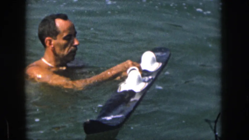 Adult man in the lake appears to position white-topped water skis before a waterskiing start in 1961 Michigan footage.