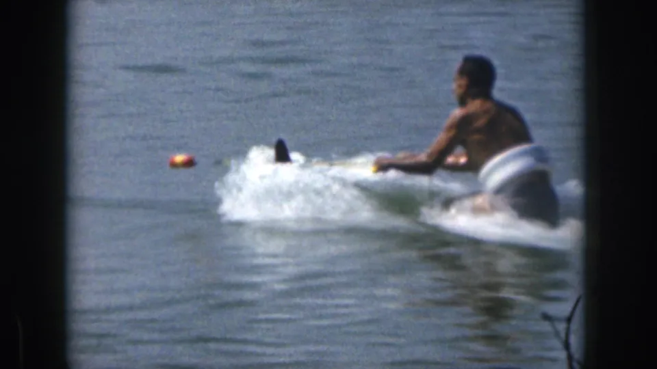 The skier starts moving behind a boat on a Michigan lake, with water spray and a tow line visible in the 1961 home movie.