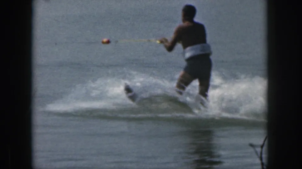 Adult man in white swim trunks balances on water skis as he is pulled across a Michigan lake in 1961 home-movie footage.