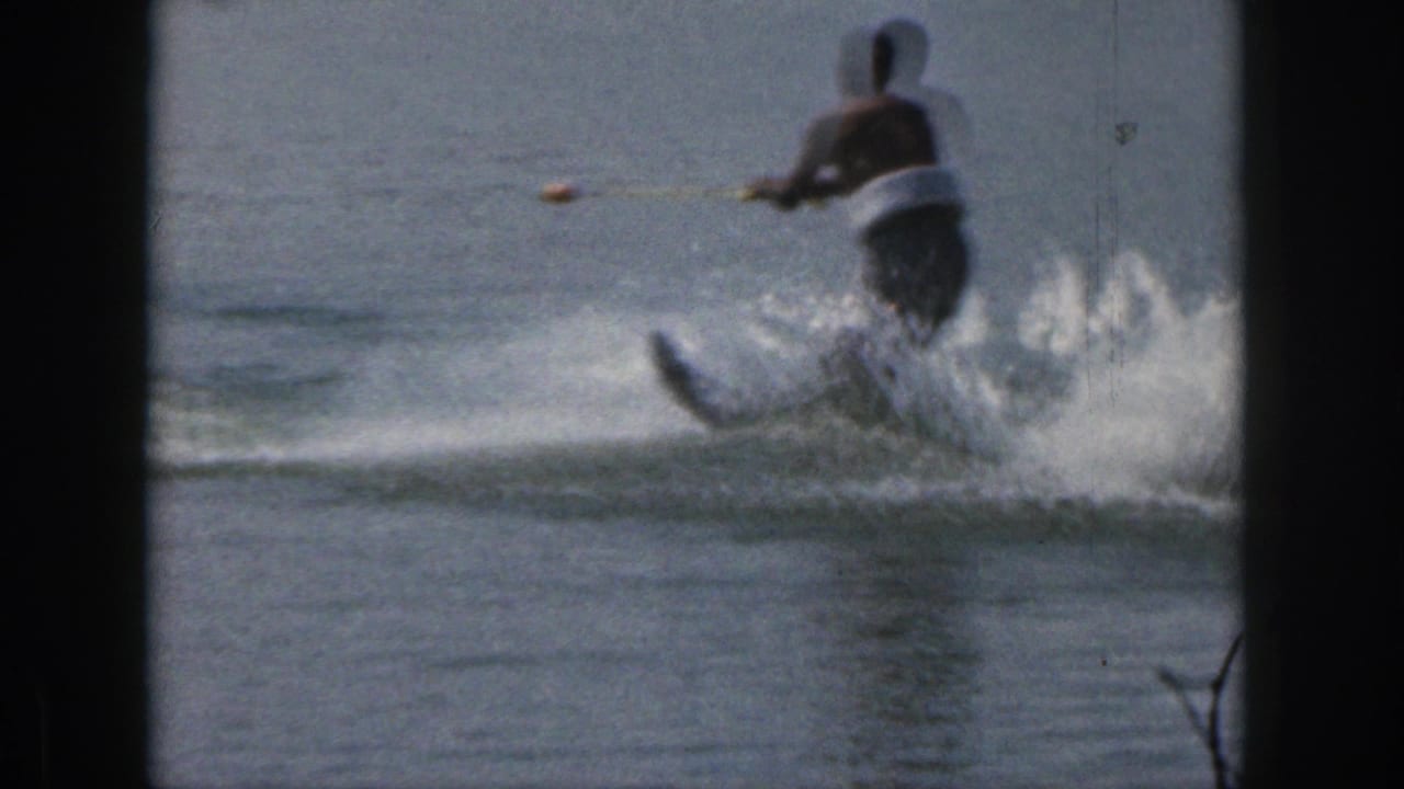 1961 Michigan home-movie frame showing a man in a life vest water skiing across a calm lake with spray behind him.