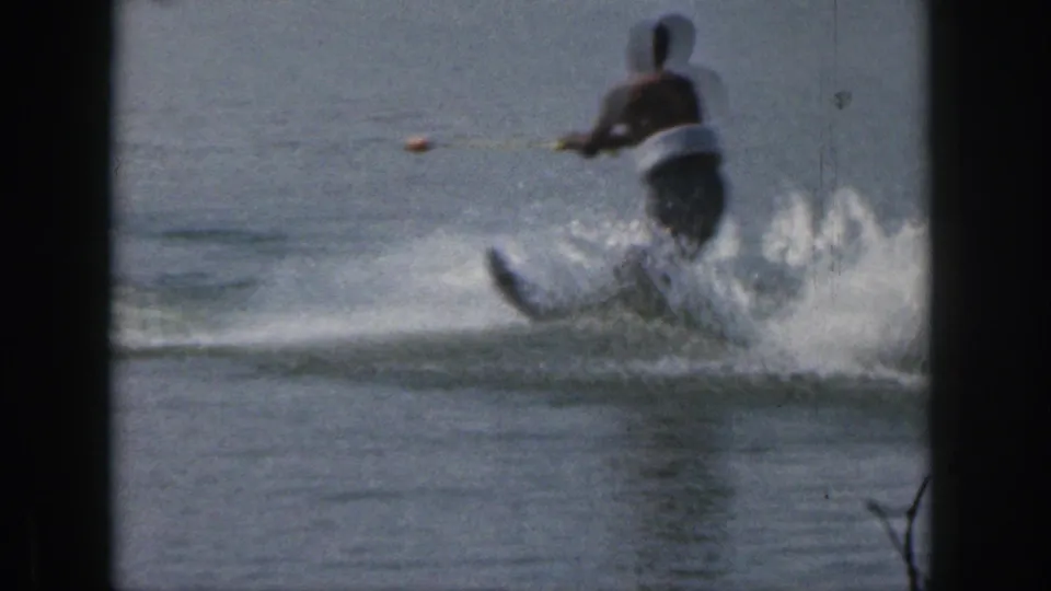 1961 Michigan home-movie frame showing a man in a life vest water skiing across a calm lake with spray behind him.