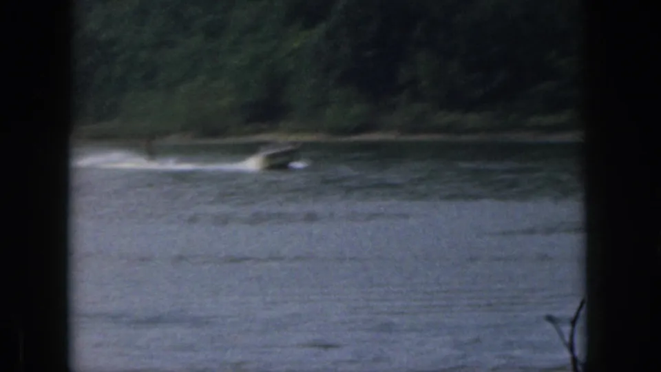1961 Michigan lake view with a small distant boat or tow wake moving near a tree-lined shoreline.