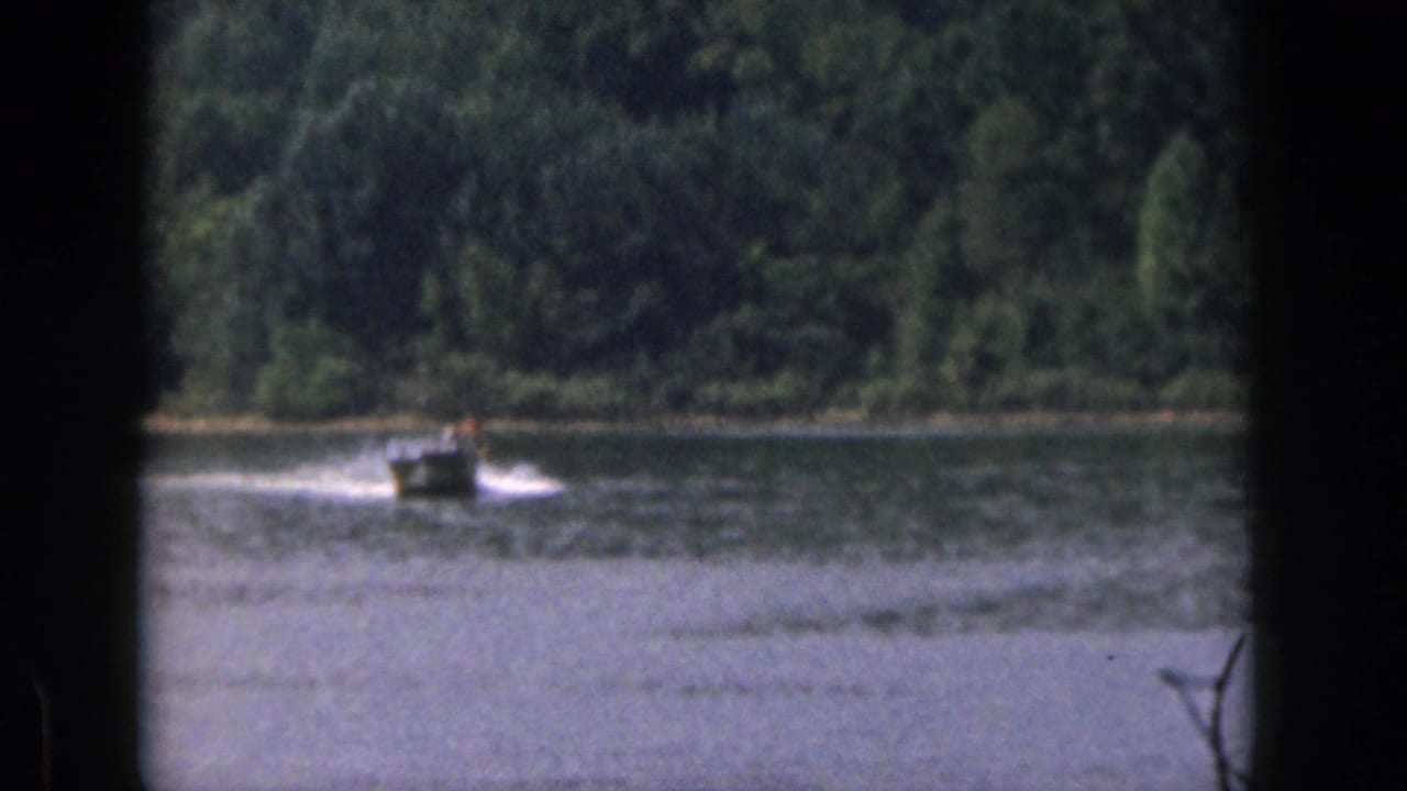 Small motorboat travels across the lake near a wooded shoreline.