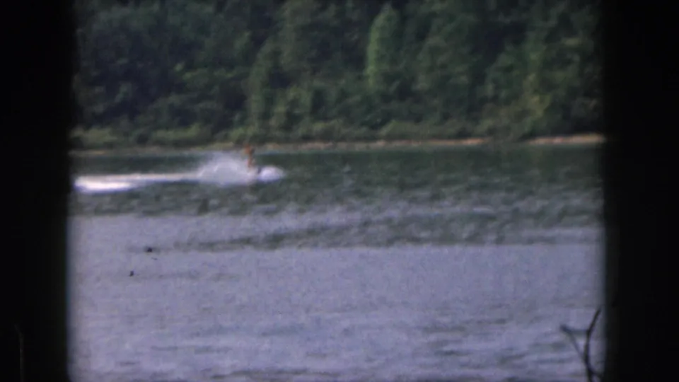 1961 Michigan lake frame with the water skier appearing at a distance behind a tow boat wake near the wooded shoreline.