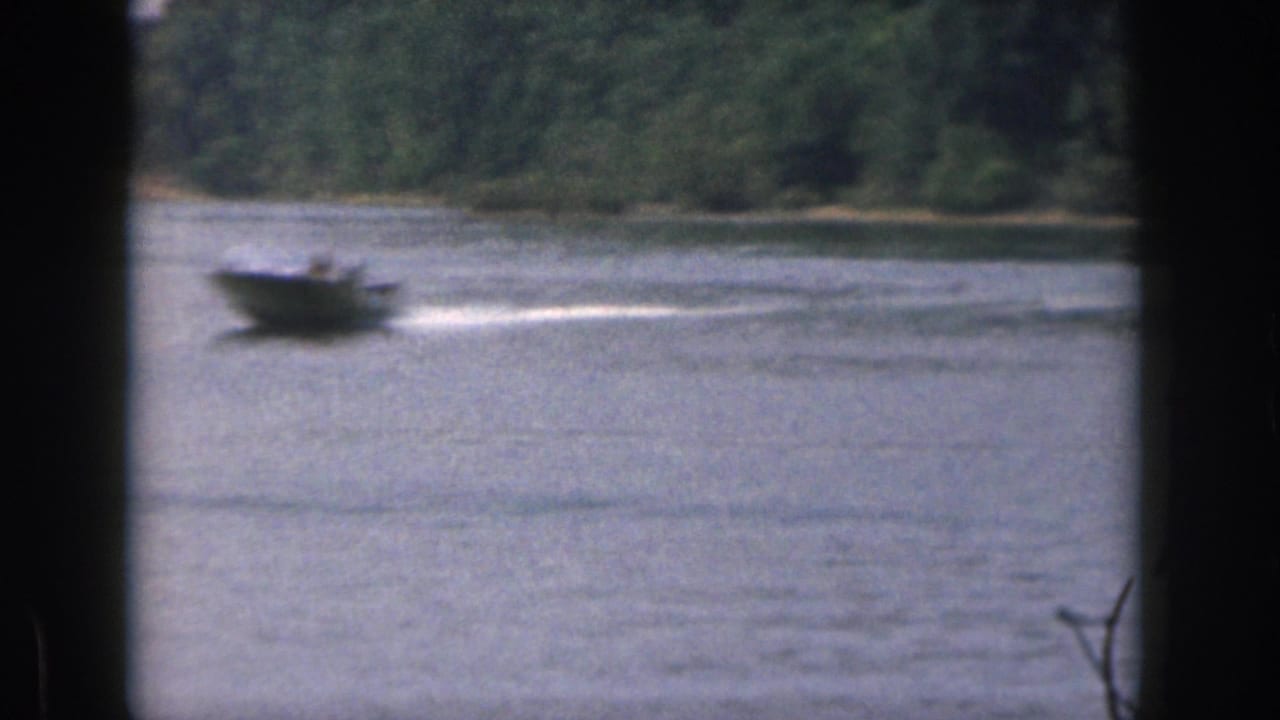 1961 Michigan home-movie frame of a motorboat moving across a lake with trees along the far shore and dark scan borders in the foreground.