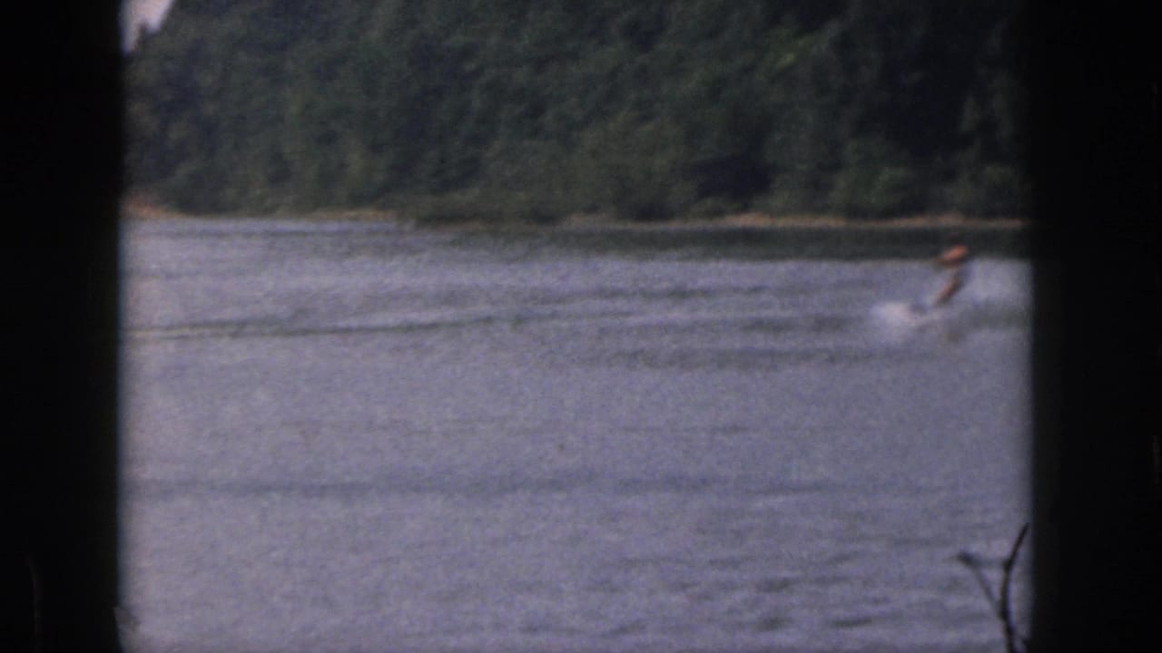Distant water skier on a Michigan lake in 1961, seen through dark frame borders with wooded shoreline behind the water.