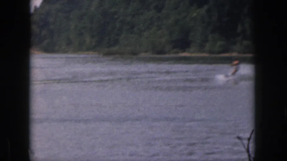 Distant water skier on a Michigan lake in 1961, seen through dark frame borders with wooded shoreline behind the water.