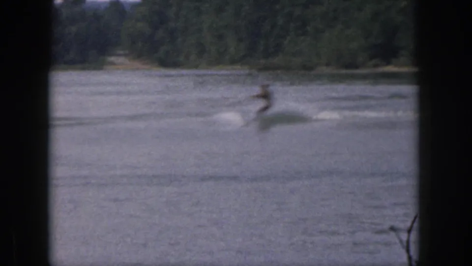 Motion-blurred 1961 Michigan water-skiing frame with the skier crossing calm lake water below a dense green shoreline.