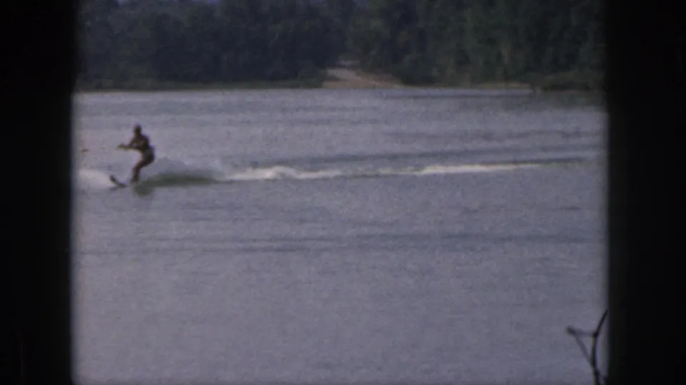 A skier in a dark swimsuit glides across a Michigan lake in 1961, leaving a bright wake against the wooded shoreline.