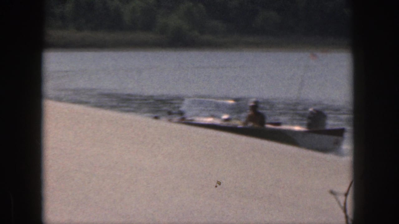 1961 Michigan home-movie frame showing a small boat with riders moving across lake water near a sandy shore.