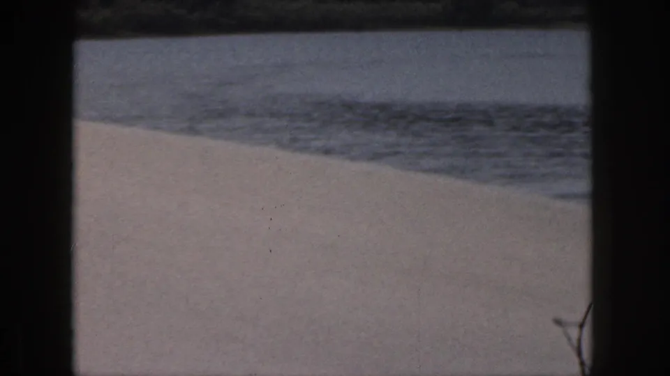 1961 Michigan home-movie shoreline frame showing calm lake water meeting a pale sandy beach.