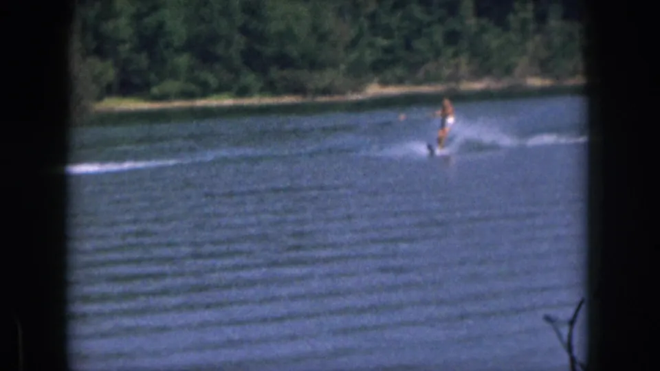 A distant water skier glides across a Michigan lake in 1961 home-movie footage, with tow spray and wooded shore visible.