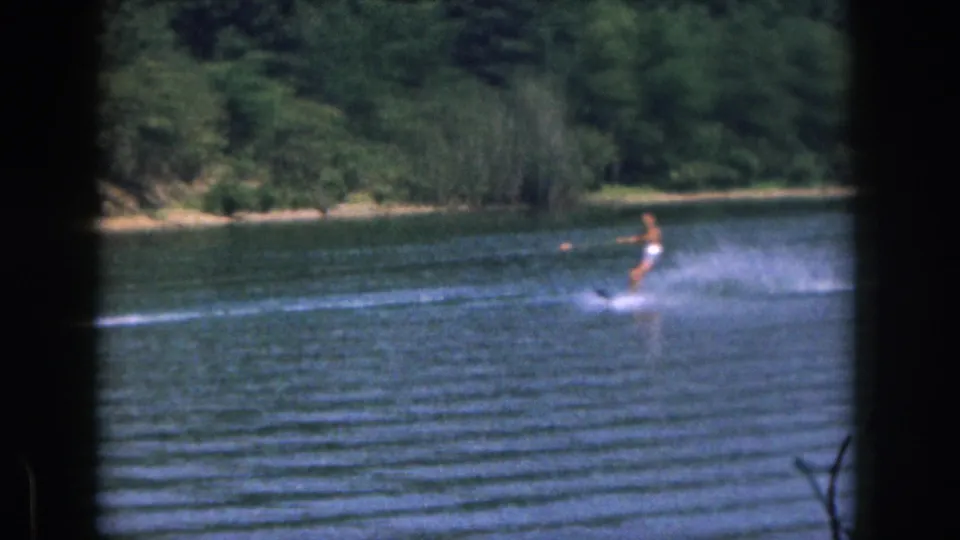 1961 Michigan lake footage shows a water skier practicing at distance, framed by 8mm borders and shoreline trees.