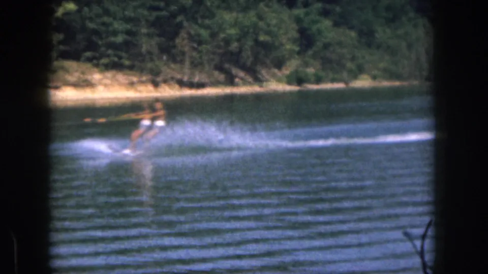 A distant skier leans while moving over a wake on a wooded Michigan lake in 1961 8mm home-movie footage.