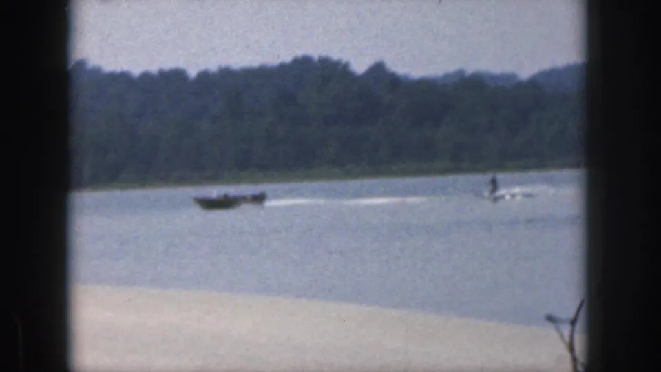 1961 Michigan 8mm frame showing a distant boat towing a water skier across open lake water with trees on the far shore.
