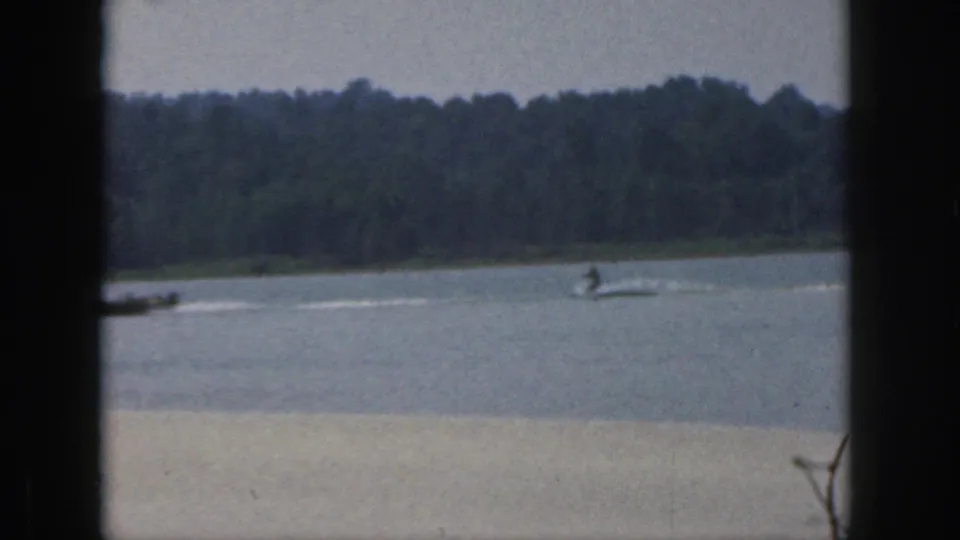 1961 Michigan home-movie frame with a boat and water skier at a distance, framed by lake water and wooded shoreline.