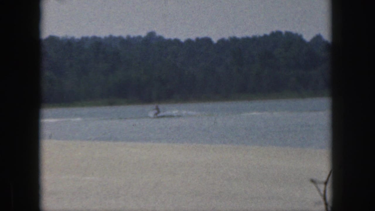 A distant water skier crosses a calm Michigan lake in 1961, framed by dark window mullions and a wooded shoreline.