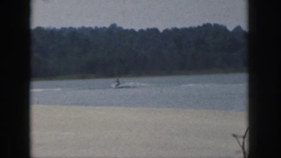 A distant water skier crosses a calm Michigan lake in 1961, framed by dark window mullions and a wooded shoreline.