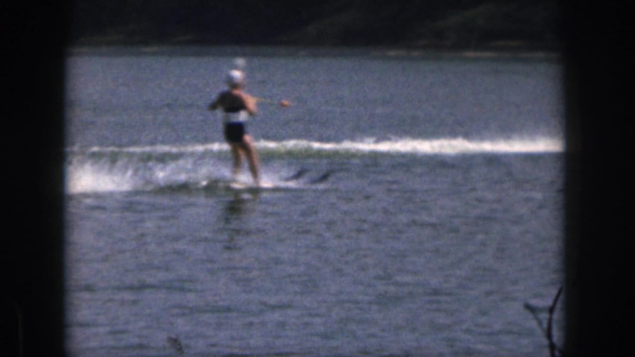 1961 Michigan 8mm frame showing a woman in a swim cap water skiing upright across a lake with a tree-lined shore behind her.