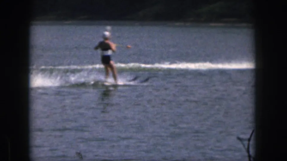1961 Michigan 8mm frame showing a woman in a swim cap water skiing upright across a lake with a tree-lined shore behind her.