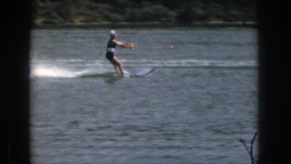 1961 Michigan 8mm still showing a woman in a dark swimsuit and white cap water skiing past rippled lake water and distant trees.