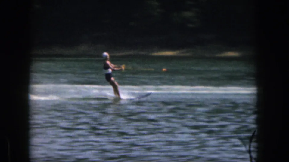 1961 Michigan home-movie frame of a woman water skiing across the lake, with a bright wake and dark tree line behind her.