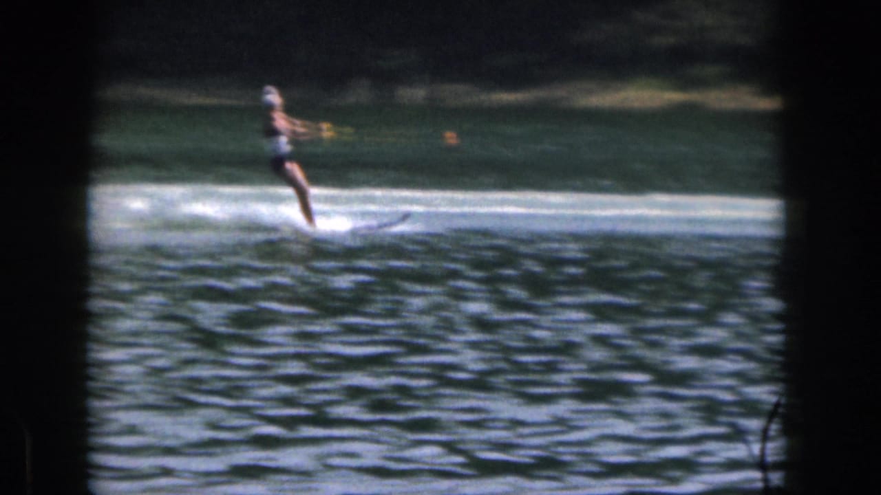 1961 Michigan 8mm frame showing a woman in a dark swimsuit and light swim cap water skiing across a tree-lined lake.
