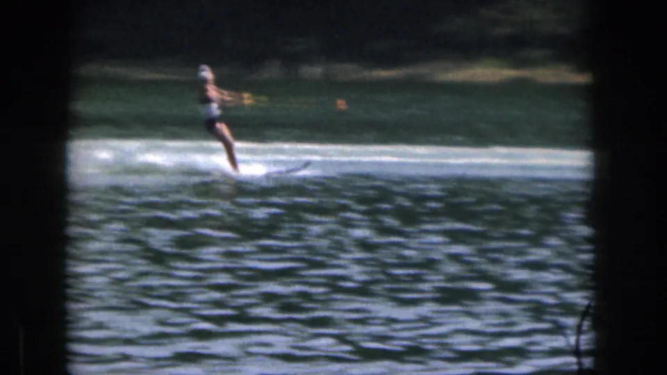 1961 Michigan 8mm frame showing a woman in a dark swimsuit and light swim cap water skiing across a tree-lined lake.