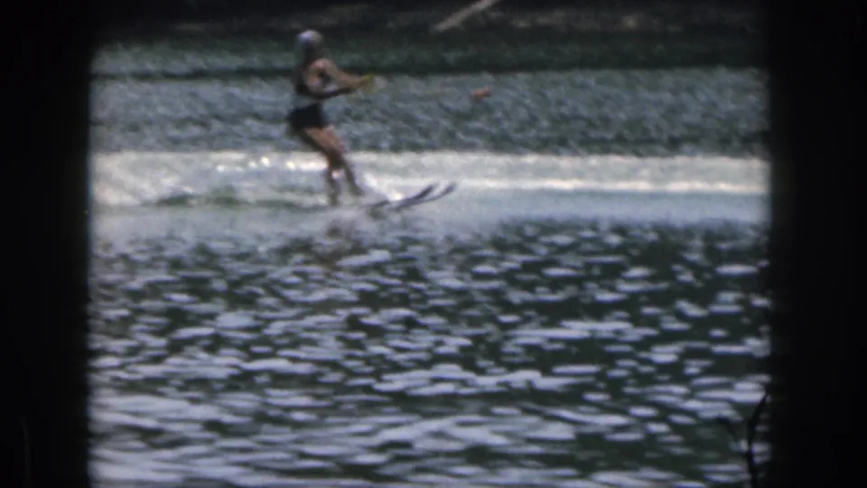 A soft 8mm frame shows the woman water skiing through bright spray, with the wooded shoreline blurred behind her.