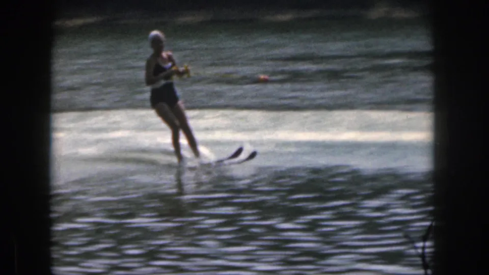 The woman in a dark swimsuit and light swim cap continues water skiing on the Michigan lake, framed by grainy 8mm borders.