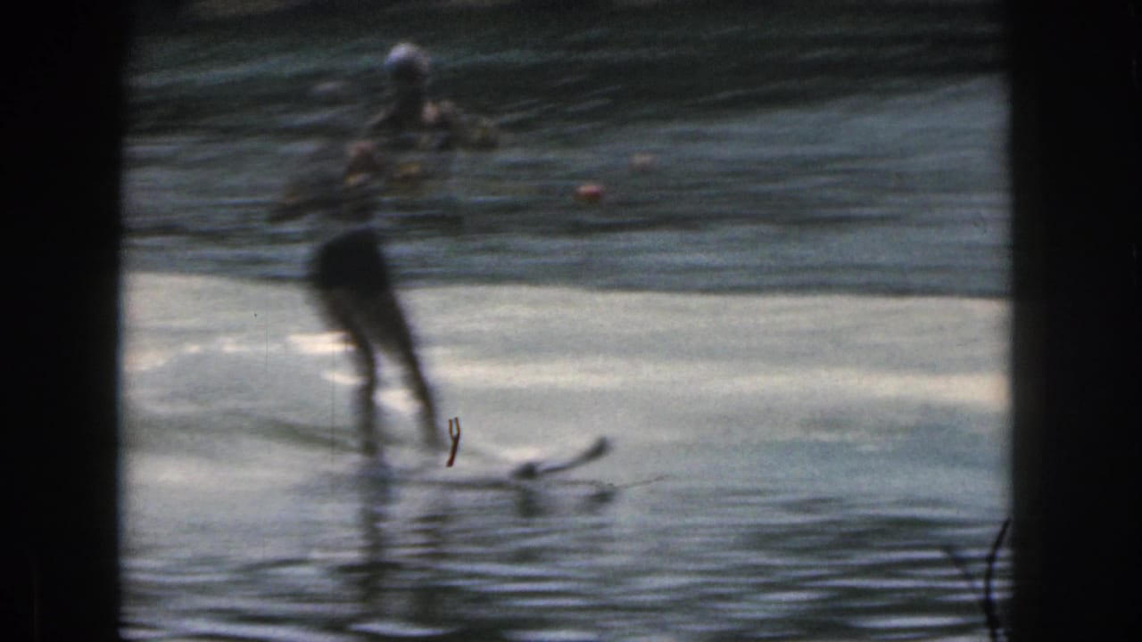 1961 Michigan home-movie frame showing a female water skier moving across a lake with a tow rope in soft focus.