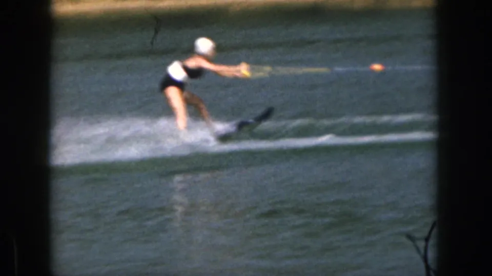 Female water skier in a dark one-piece swimsuit and white swim cap skims across a Michigan lake in 1961 footage.