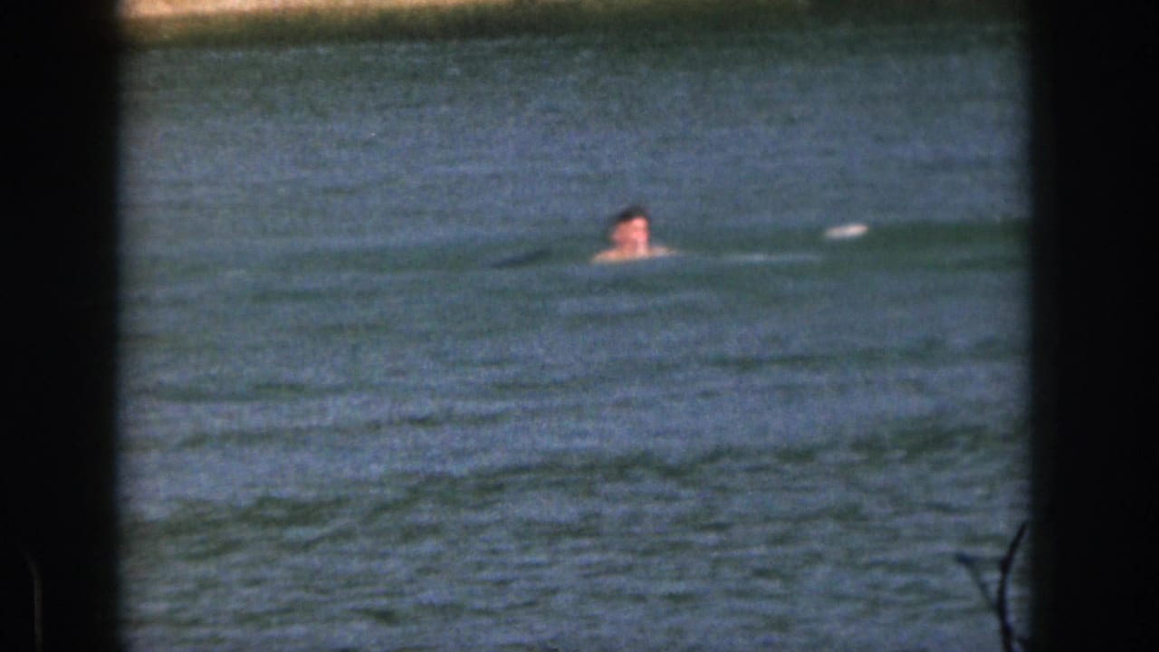 1961 Michigan 8mm frame showing a distant swimmer in calm blue water, seen through dark frame edges.