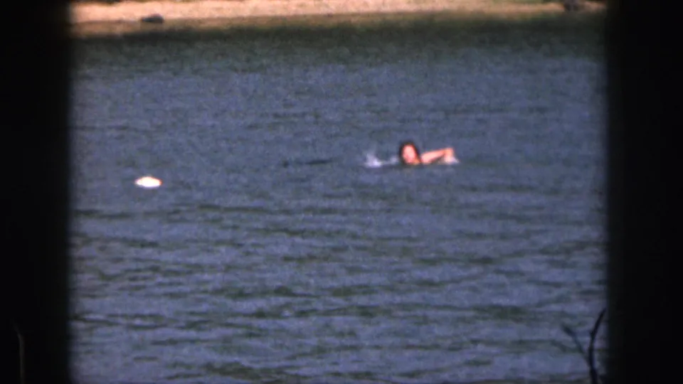 Distant 1961 Michigan 8mm view of a swimmer in blue water beside a small bright object or reflection.