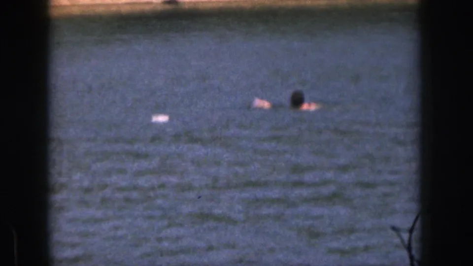1961 Michigan home-movie frame showing a distant swimmer from behind in calm blue water with visible film grain.