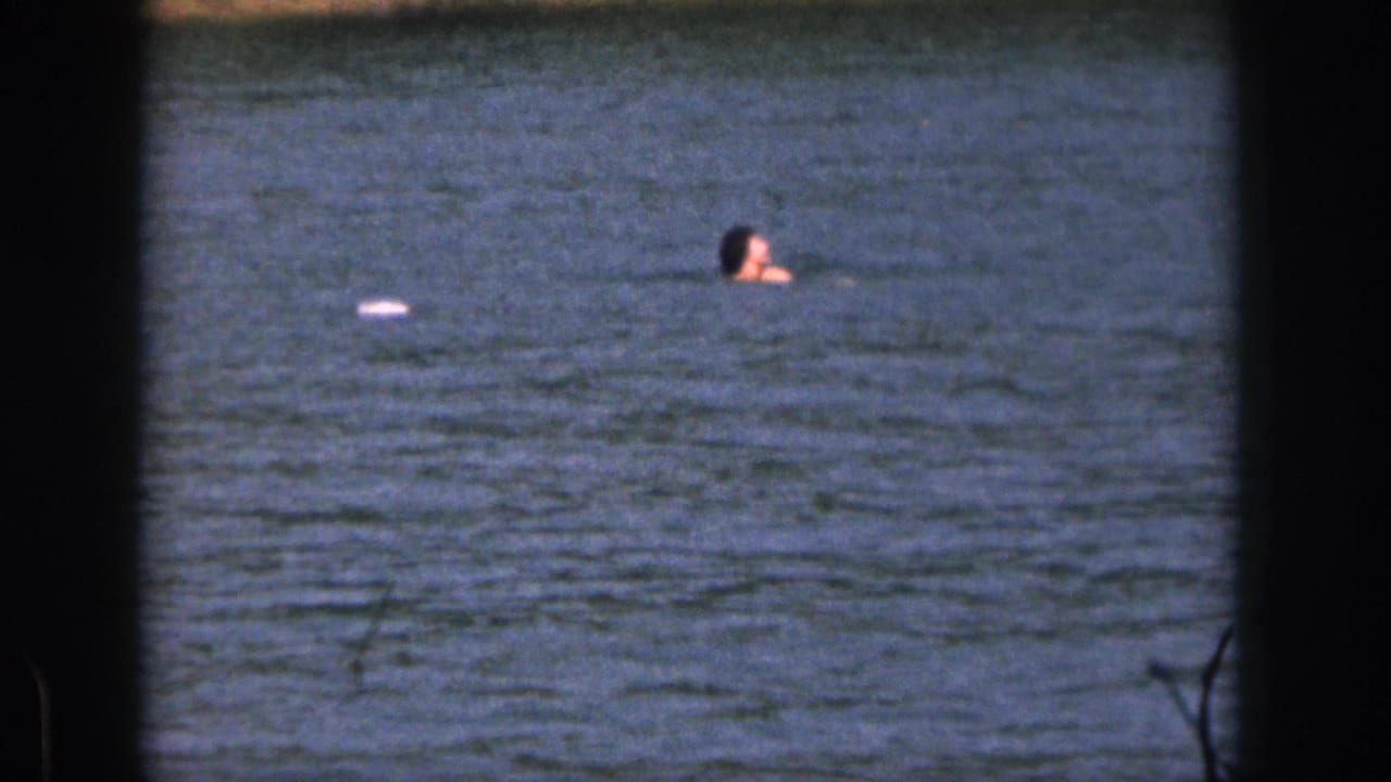 1961 Michigan 8mm frame showing a person swimming in a lake, framed by dark film borders with shoreline visible beyond the water.