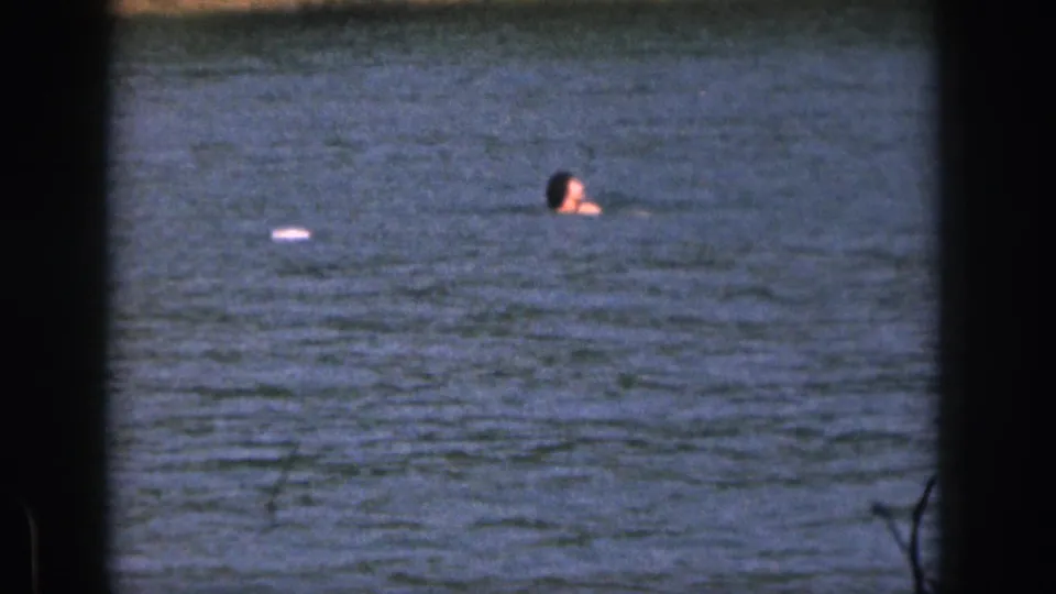 1961 Michigan 8mm frame showing a person swimming in a lake, framed by dark film borders with shoreline visible beyond the water.
