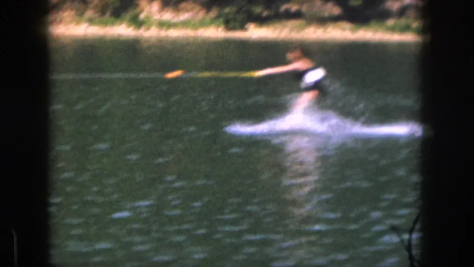 1961 Michigan home-movie frame showing a water skier in a dark and white swimsuit crossing the lake with tow rope and spray visible.