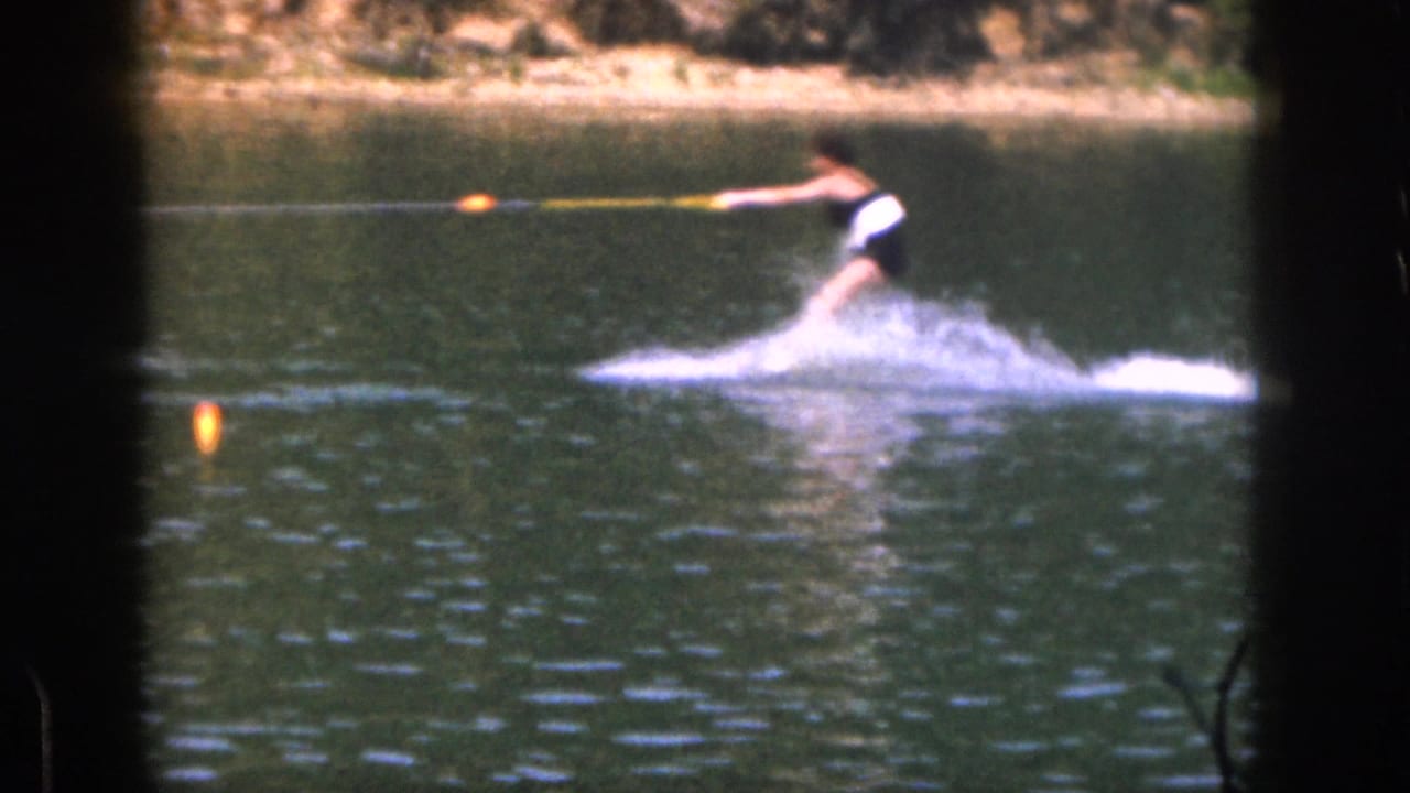 1961 Michigan 8mm frame showing a woman in a one-piece swimsuit water skiing across a lake near a wooded shoreline.