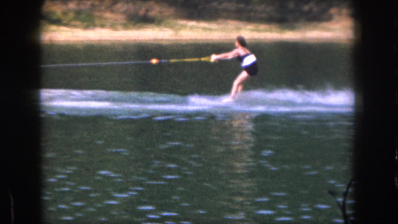 A woman water skis on a Michigan lake in 1961, leaning back as spray trails behind her in soft 8mm color footage.