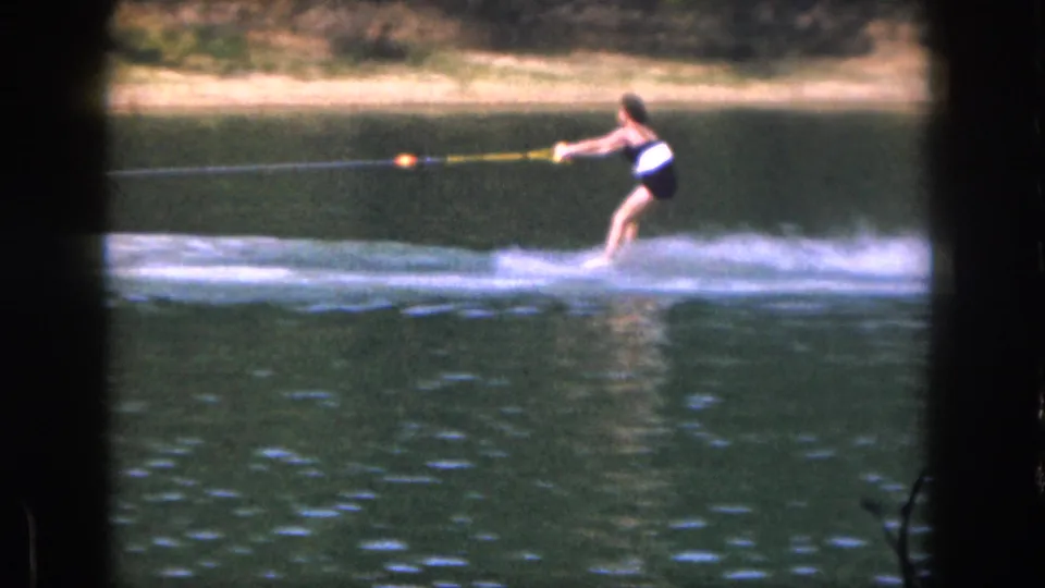 A woman water skis on a Michigan lake in 1961, leaning back as spray trails behind her in soft 8mm color footage.
