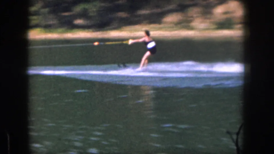 1961 Michigan home-movie frame of a woman water skiing past the camera, with broad white wake and shoreline in the background.