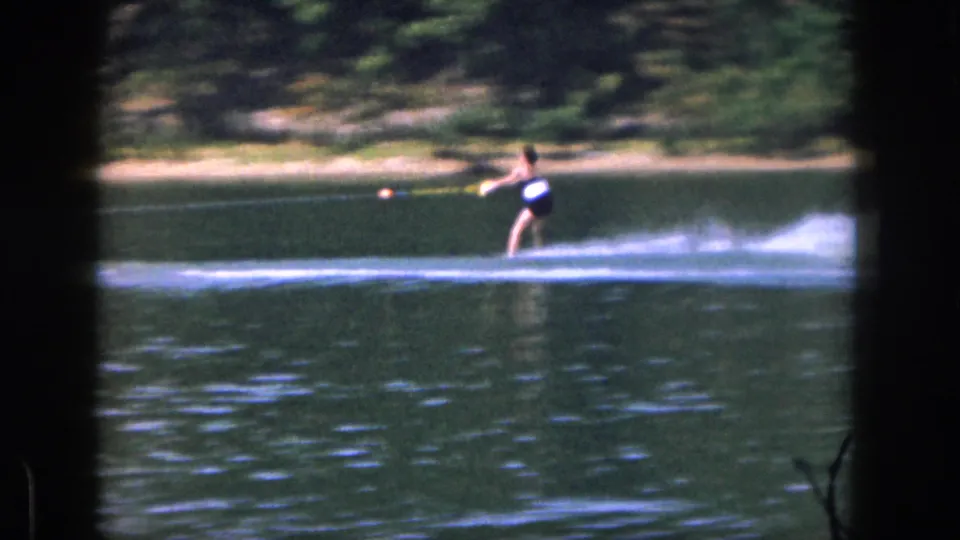 A distant 1961 Michigan 8mm view shows a woman water skiing across green lake water, framed by dark film edges and a wooded shore.