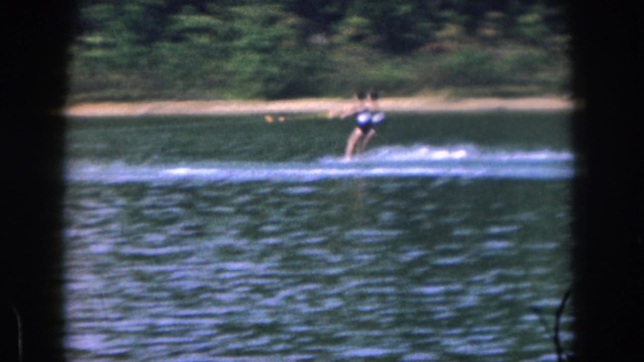 1961 Michigan home-movie frame showing a distant water skier on a calm lake, framed by dark foreground borders.