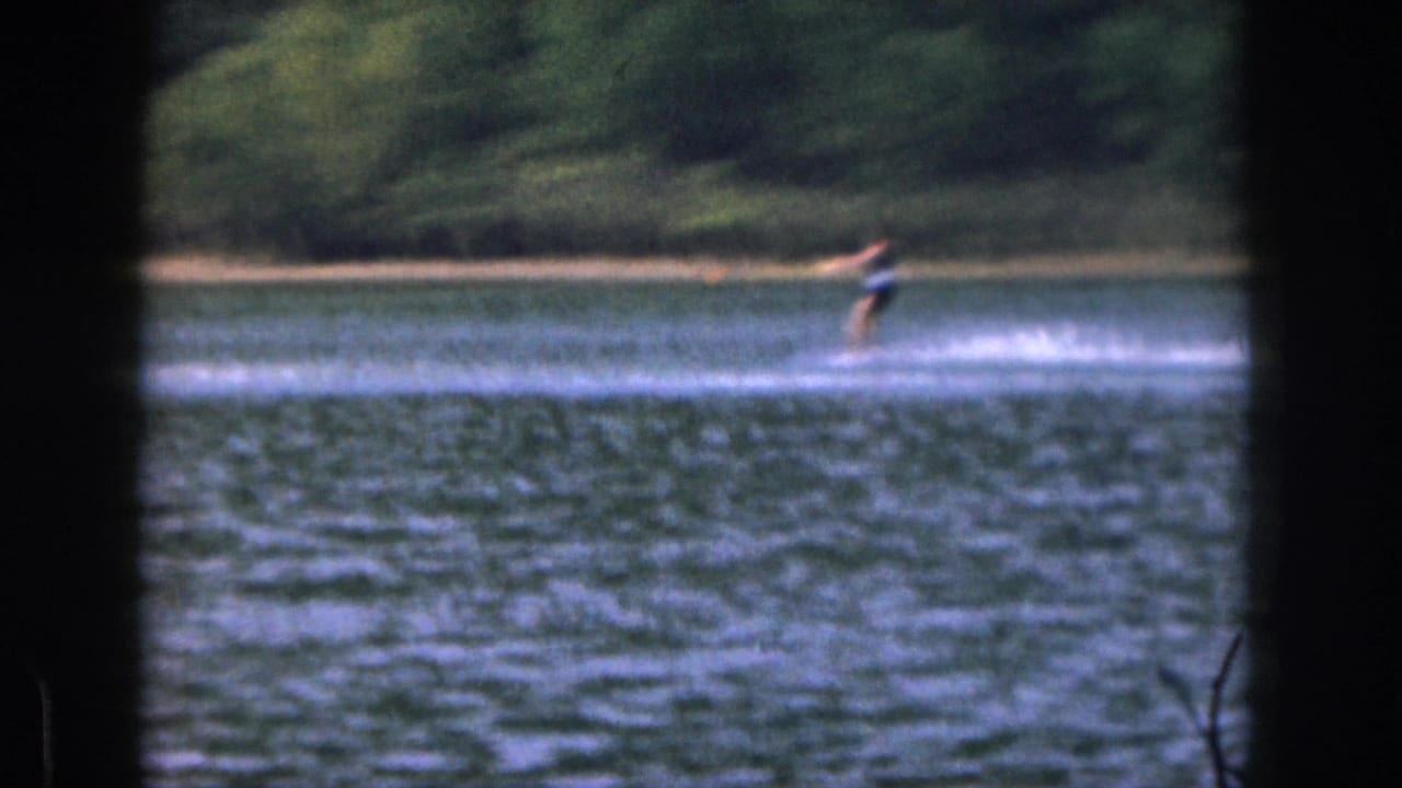 Distant skier in dark swimwear moves across a Michigan lake in soft 8mm footage from 1961.