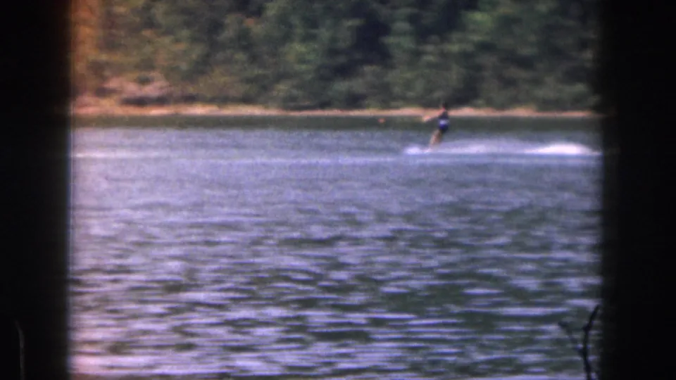 Soft 8mm frame of a water skier crossing a Michigan lake in 1961, with trees and shoreline behind.